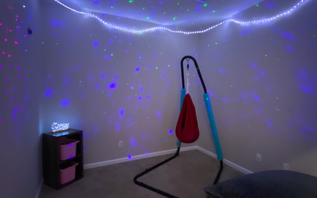 Darkened sensory room with lights surrounding the top of the walls and projected onto the ceiling, with a hammock swing in one corner and an oversized pillow in another corner