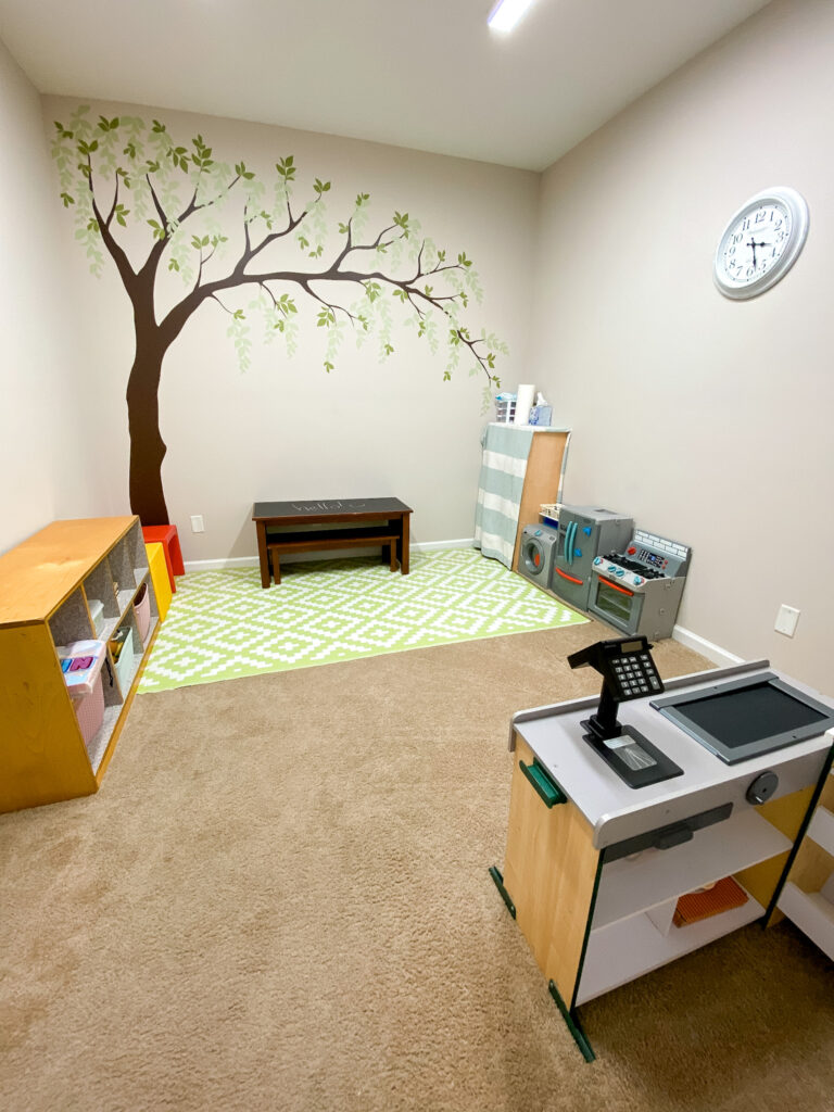 Pretend play room with large tree on the back wall, with grocery store, kitchen set, and other toys for kids of all ages