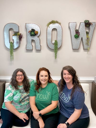 Co-owners Christy Clutter, BS, Ed., COTA/L and Victorie Steed, MS, OTR/L sit on either side of their employee, Jenny Newton, COTA/L in their waiting area in front of their decorative "GROW" sign